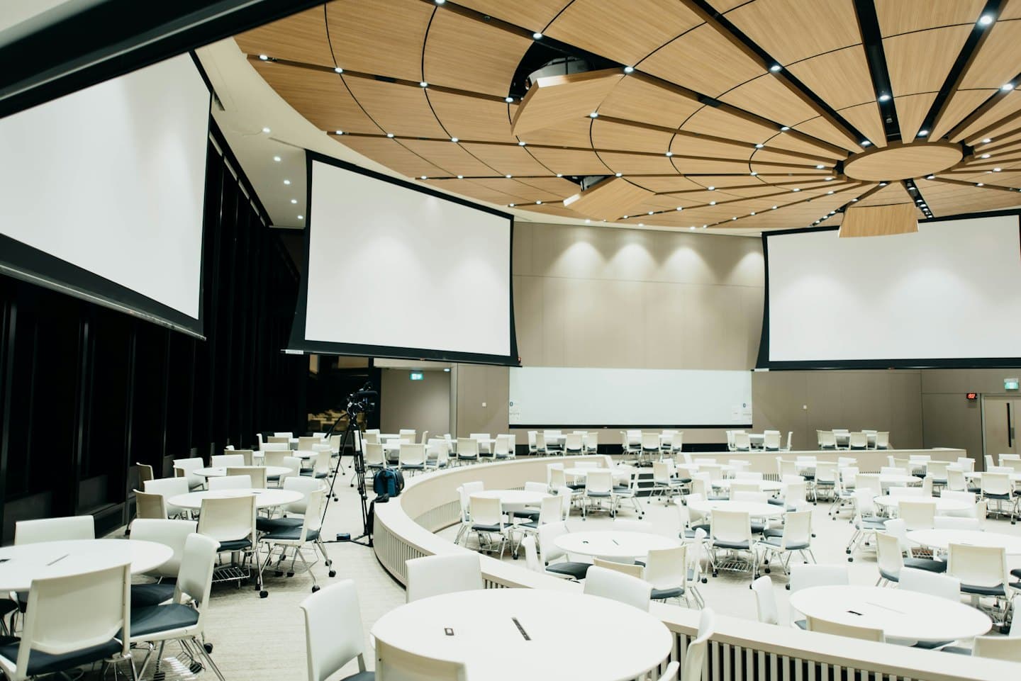Leerer moderner Konferenzsaal mit runden Tischen vor Projektionsleinwänden.